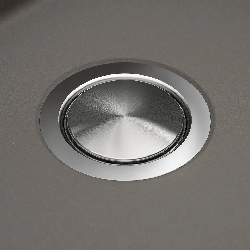 Stainless steel sink drain cover, smooth and polished, embedded in dark quartz countertop.