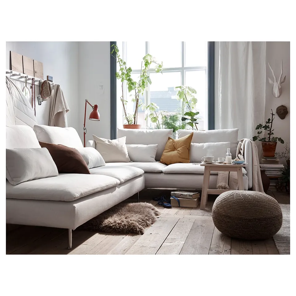 Cosy living room with white l-shaped sofa, colourful pillows, wood floor, and large window. Decorated with plants and a unique lamp.
