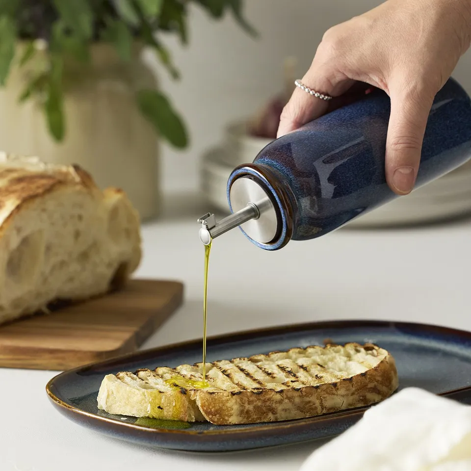 Person drizzles oil onto grilled bread from a blue BRUGDHAJ bottle with a silicone seal and spout cover.