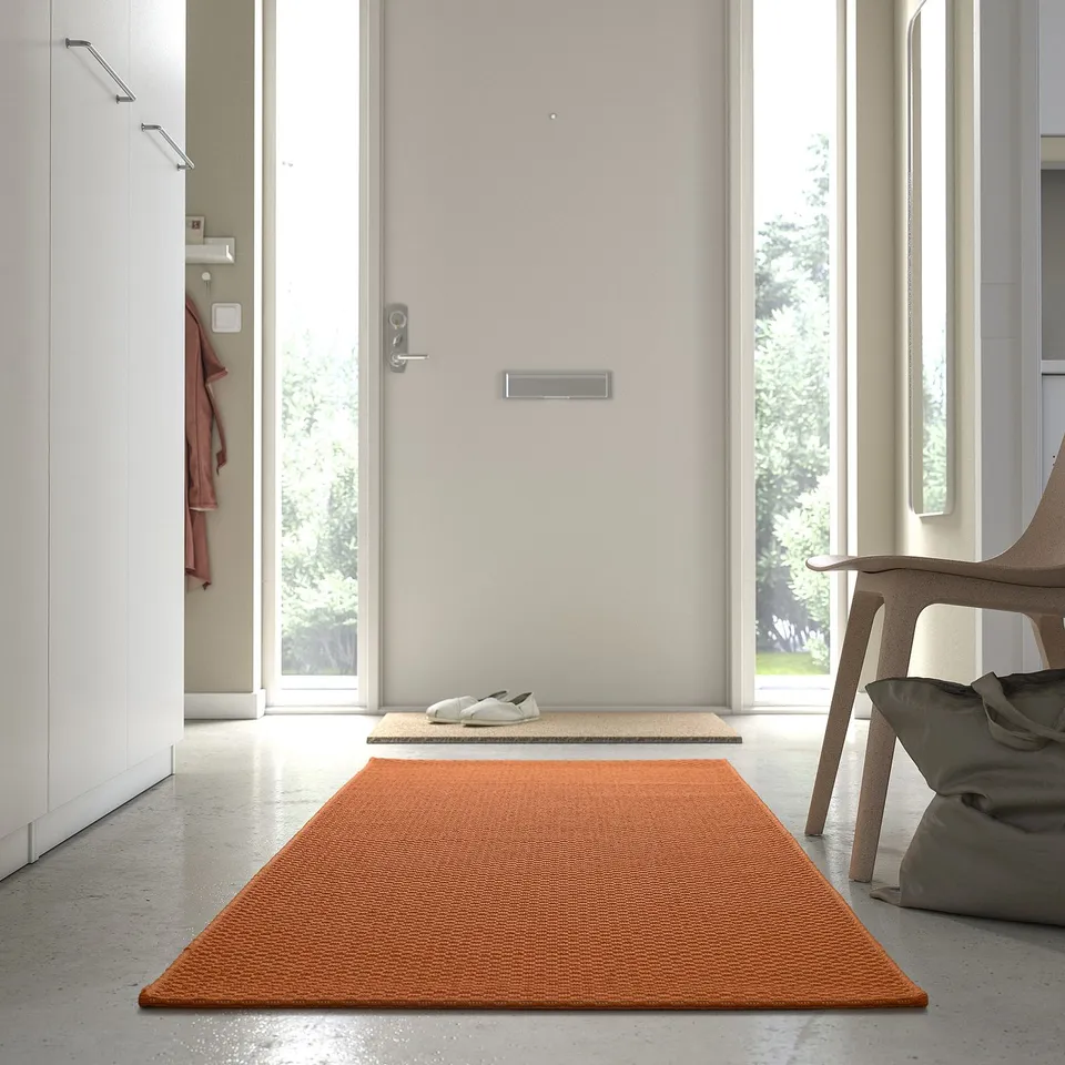 Orange rug in modern entryway next to grey chair and closet with red jacket.