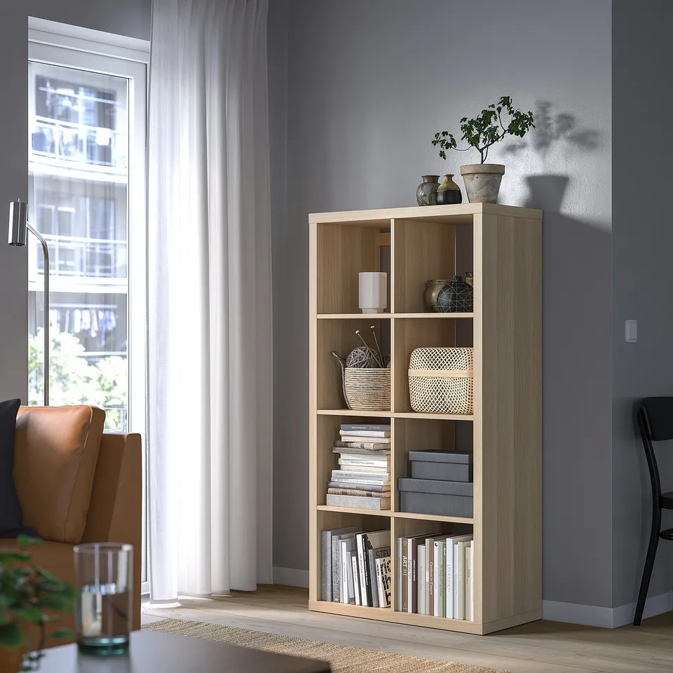 KALLAX shelving in light wood holds books, baskets, and decor between window and chair.