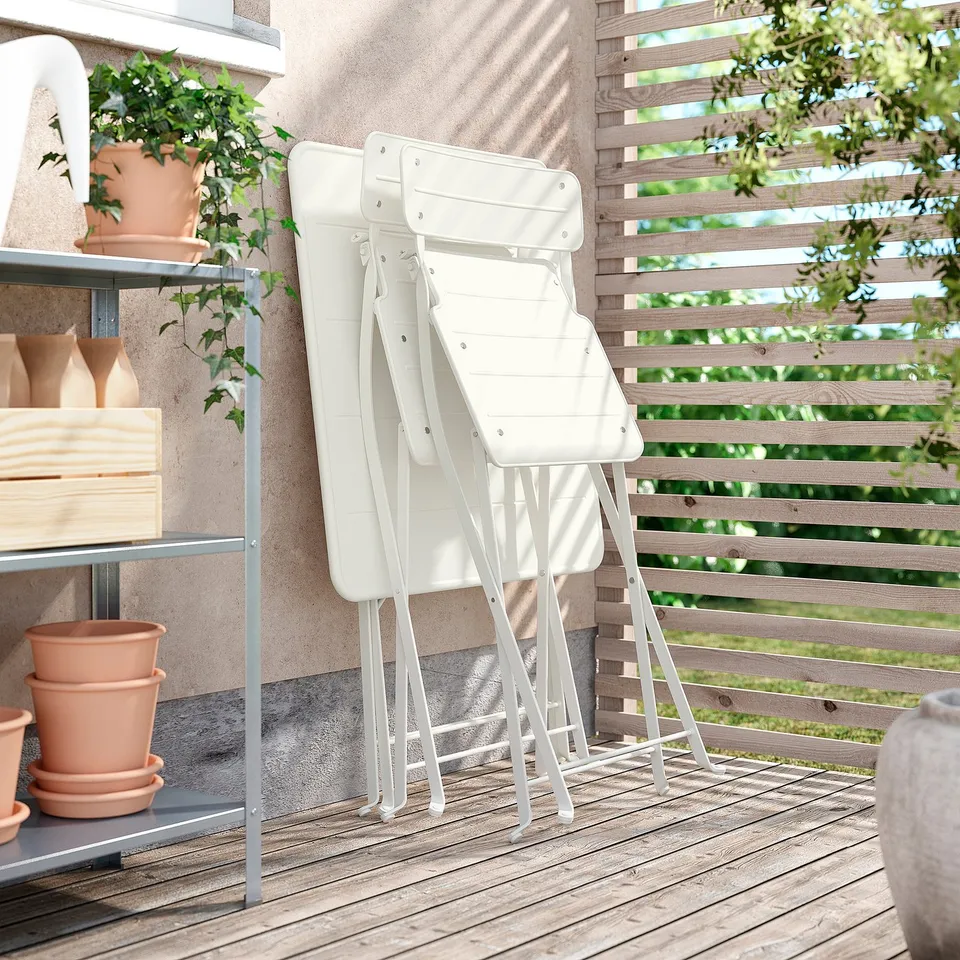 Two white folding chairs stacked against a rustic wall on a wooden deck, surrounded by potted plants.
