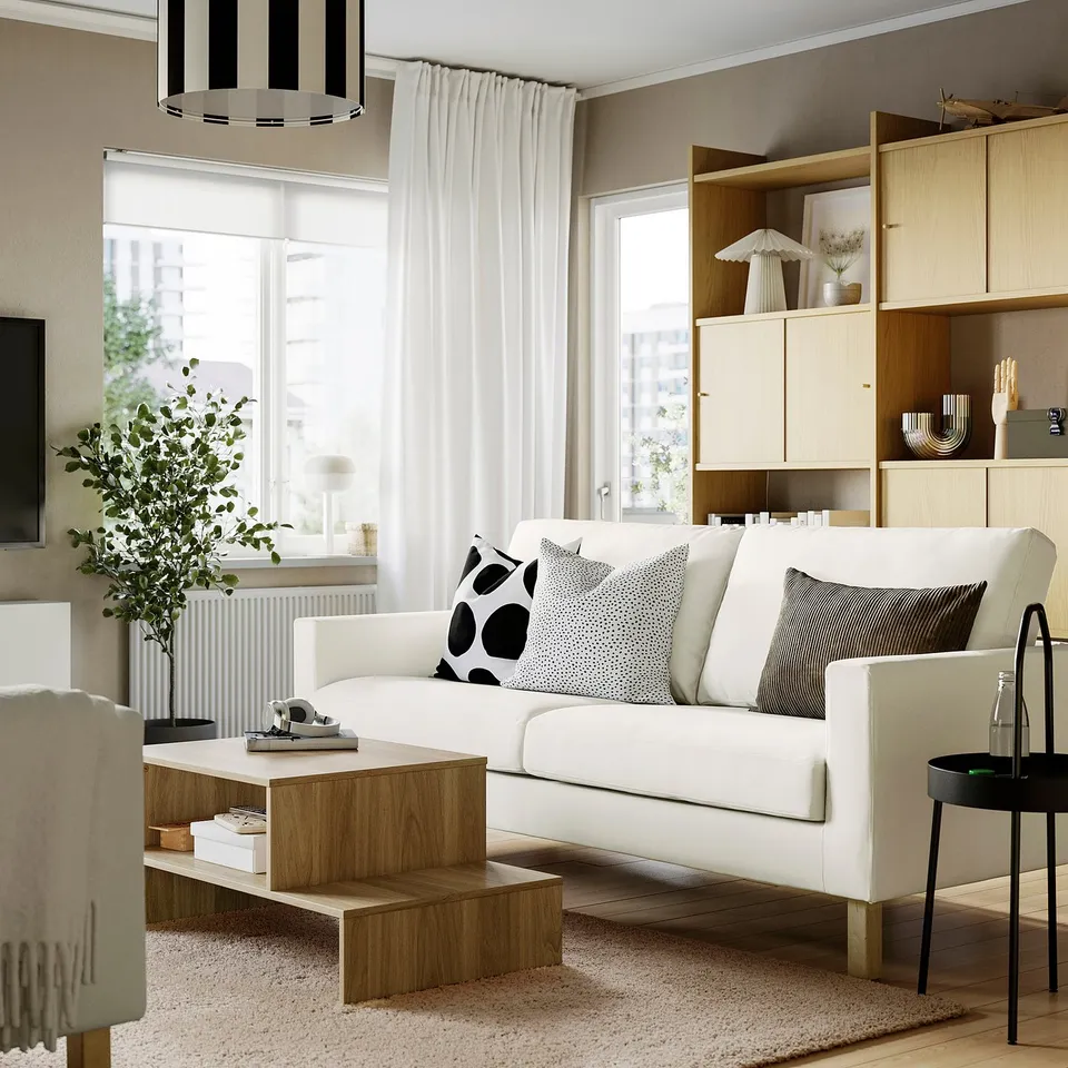 Modern living room with white sofa, pillows, coffee table, bookshelf, window curtains, side table, and rug.