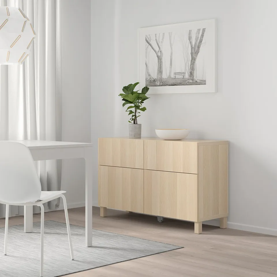 Bright room with white chair, light wood cabinet, plant, bowl, artwork; minimalist design.