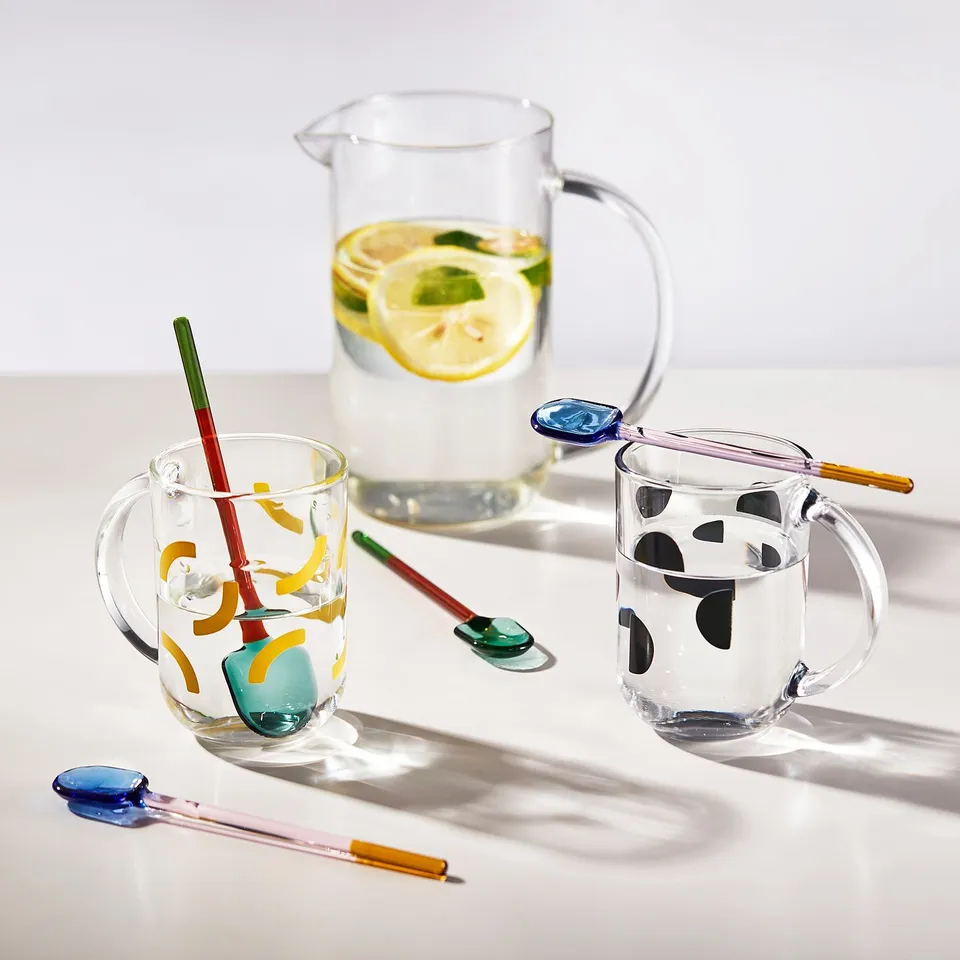 Three colourful glass mugs with abstract designs and handles, each holding lemon slices and water, with matching spoons.