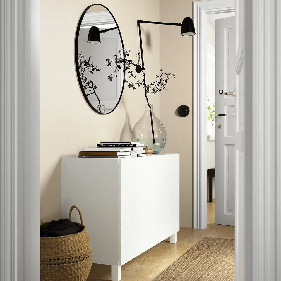 A minimalist entryway with a white LAXVIKEN door on the right, featuring a round mirror above a console with decor items.