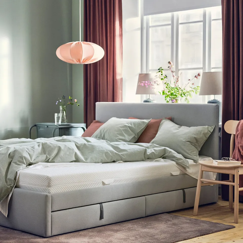 Cosy bedroom with grey mattress, pillows, and hanging lamp. Windows let in light, with a wooden chair and rug.