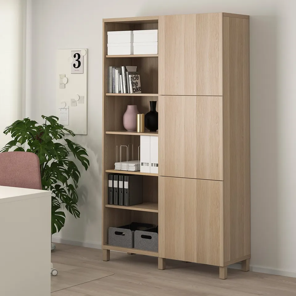 Light wood BESTÅ cabinet with open and closed storage, displaying books, vases, and boxes. It stands taller than nearby house plant and desk.