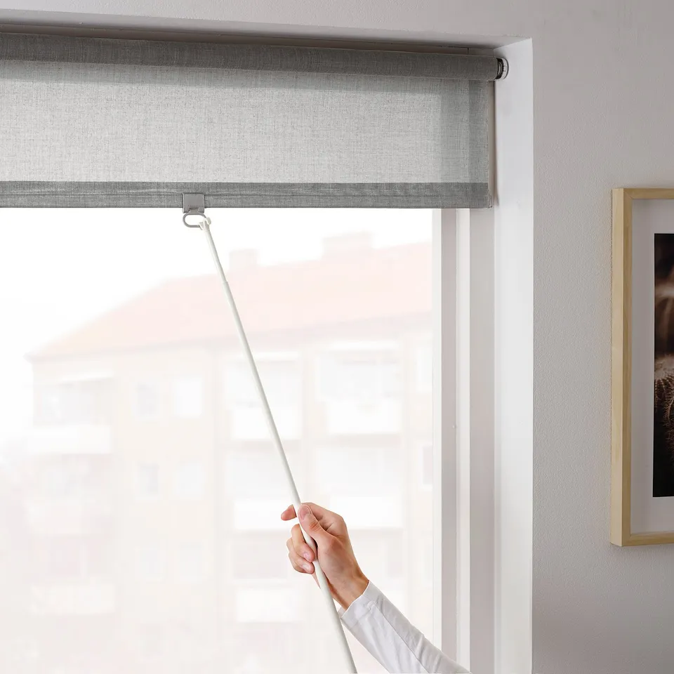 A hand pulls a grey roller blind down, demonstrating its light-filtering and privacy. The cordless design adds safety.