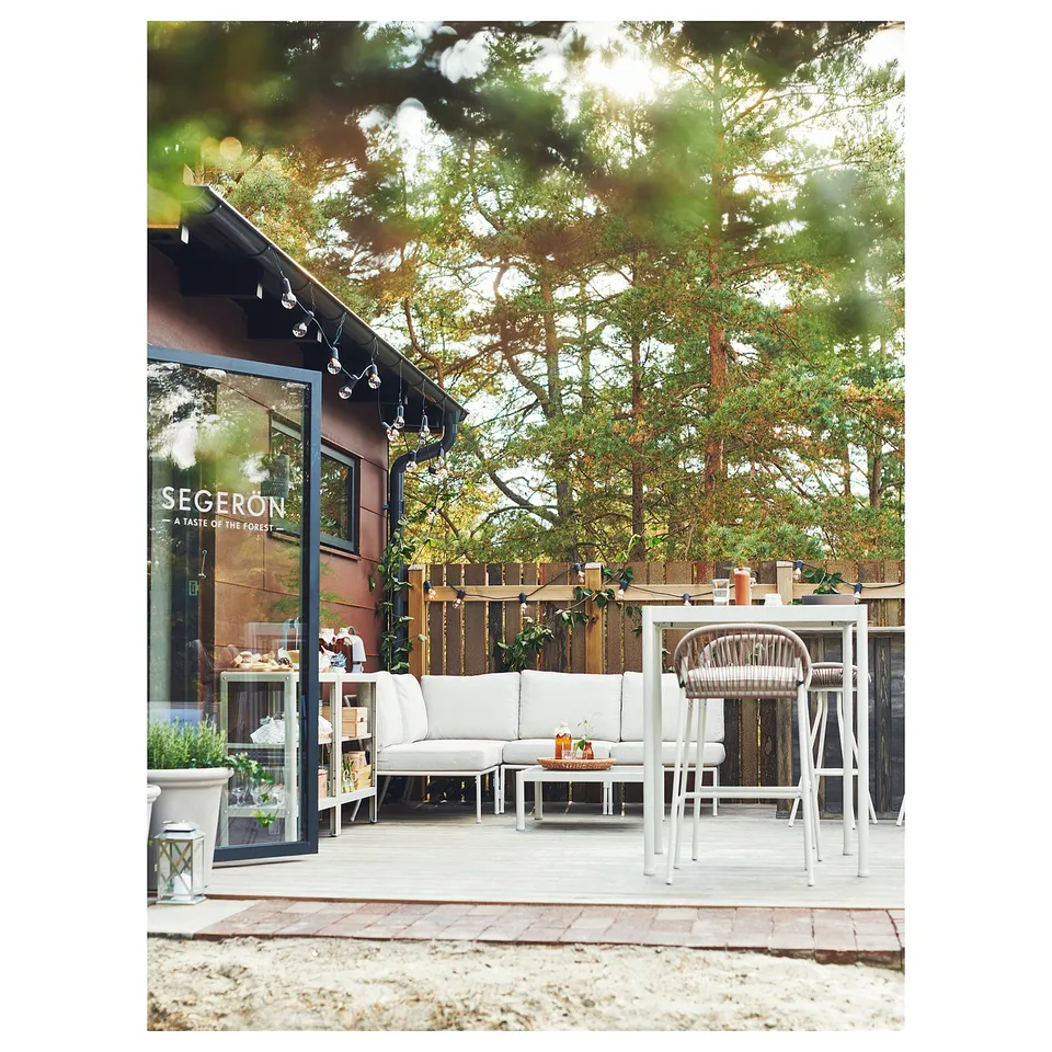 Modern outdoor patio with white SEGERÖN furniture, including a sofa and high table, set against a wooden building with string lights.
