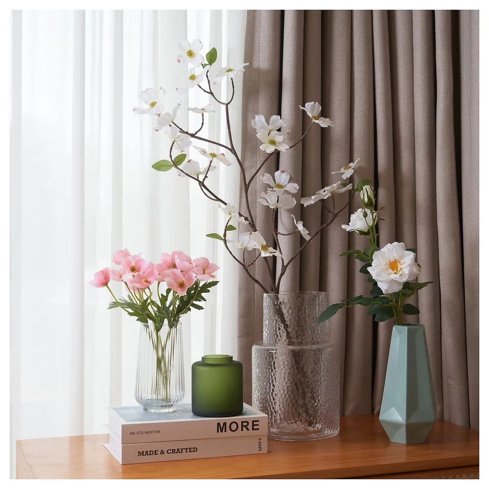 Three vases with flowers: one clear, one green, one blue. Flowers in pink and white. On a wooden table with books, curtains in background.