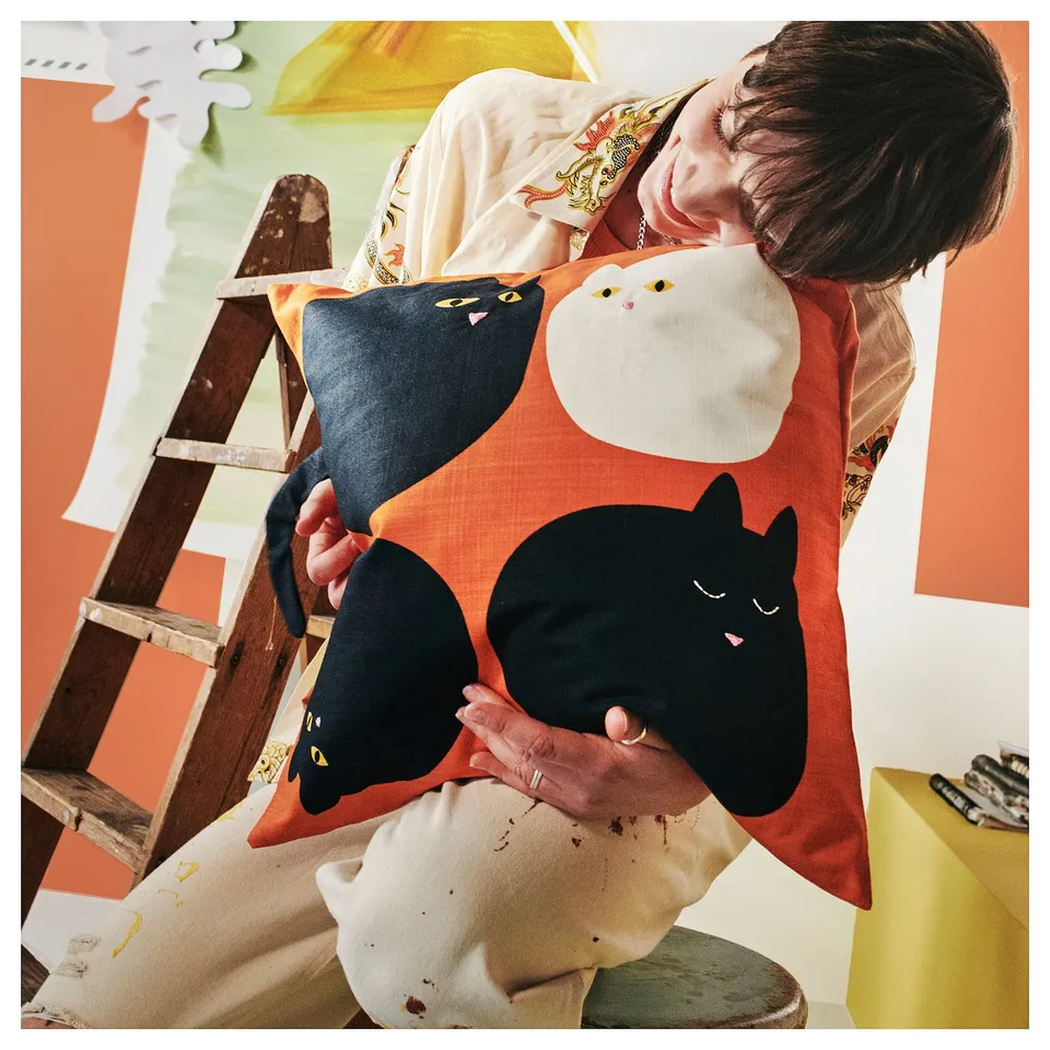 A person holds a playful cat-shaped cushion cover in orange with embroidered black and white cats.
