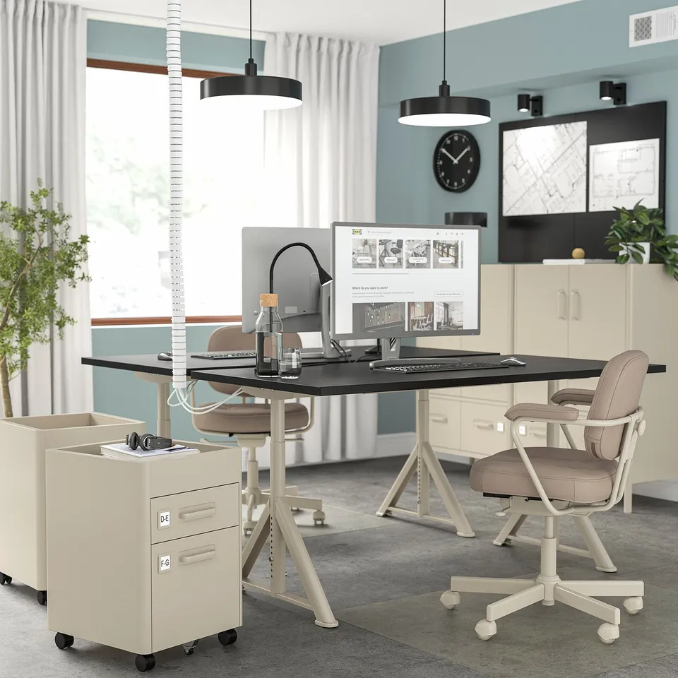 Modern home office with IDÅSEN beige desk, adjustable height, and cable management. Beige chair, cream cabinetry, light blue walls, & stapler lamp.