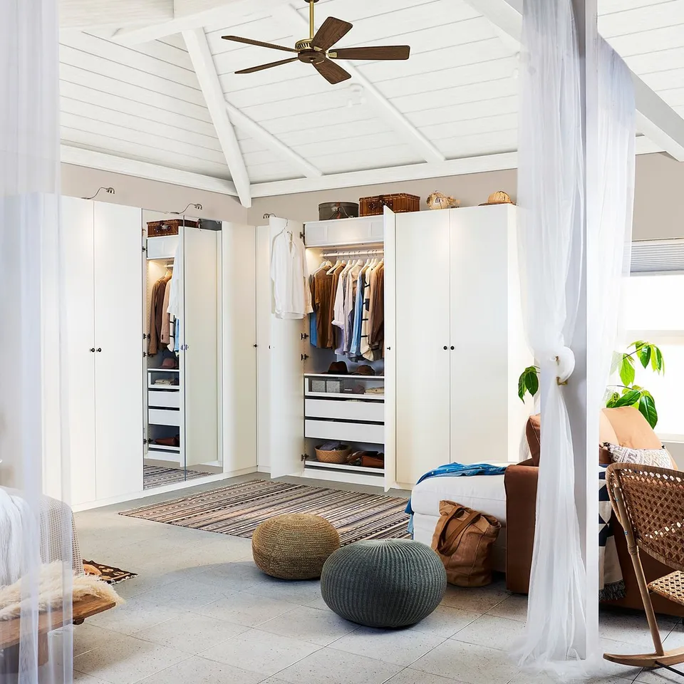 Bright, white closet with mirrored door, next to bed in airy room, light wooden floor, two round pouffes at foot.