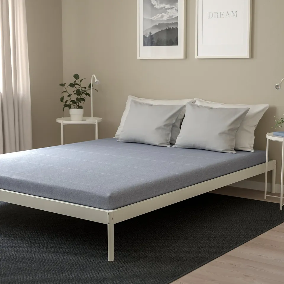 Modern bedroom with grey mattress, white bed, two nightstands, lamps, plants, wall art, dark rug, light curtains.