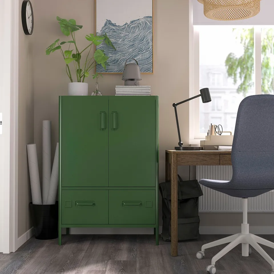 Modern office with green IDÅSEN cabinet, grey chair, wooden desk by window, plant, lamp, and artwork.