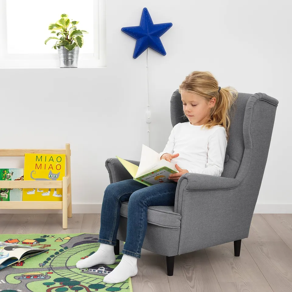 Girl reads in grey STRANDMON chair on rug, near books, star decor, and plant.