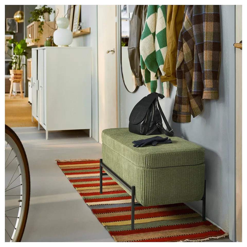 Green bench with cushion & storage in hallway, coats & a bag beside, colourful rug under.