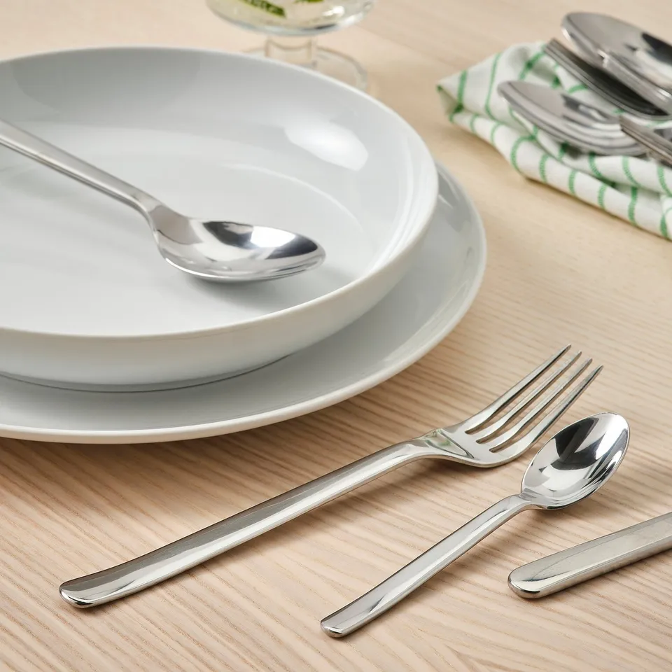 Stacked white dishes with polished silver fork, spoon, and teaspoon arranged neatly on wooden table.