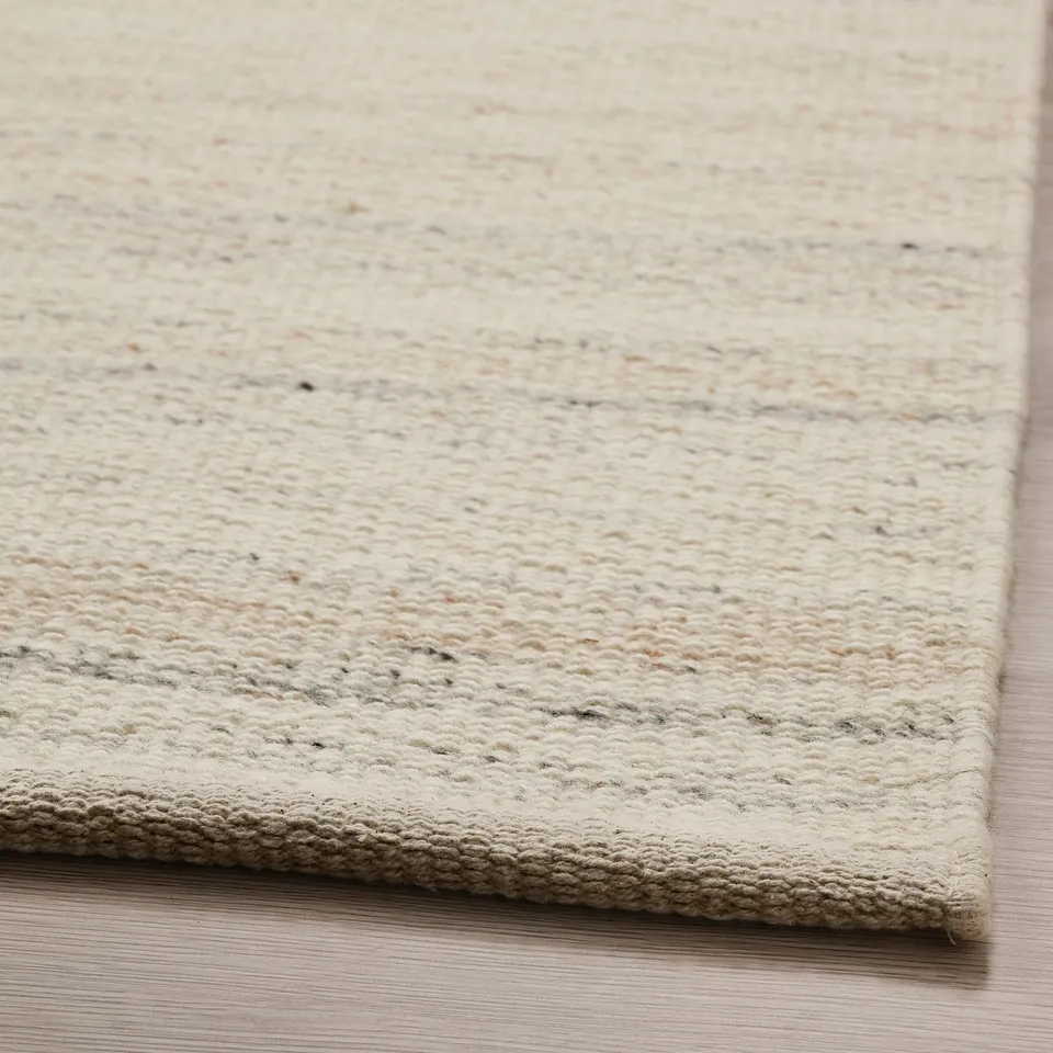 Light beige, hand-woven rug displayed on a wooden floor. Soft, textured wool surface visible, highlighting its quality and comfort.