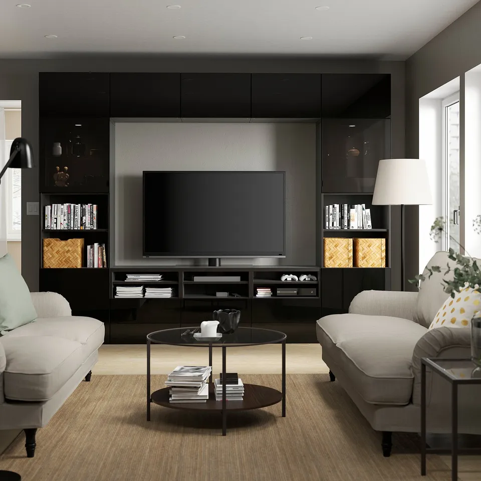 Black BESTÅ tv stand with glass doors and adjustable shelves between two windows, flanked by grey sofas.