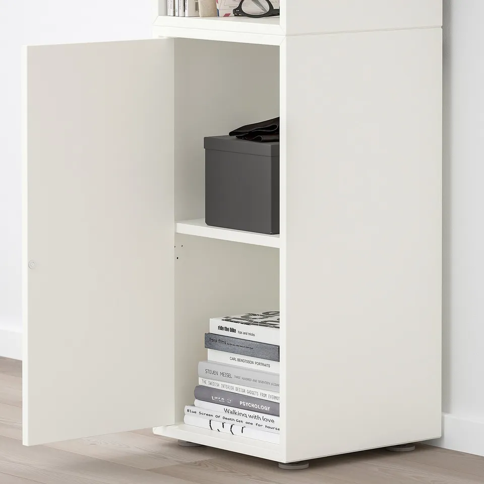 White EKET shelf unit with open shelves. Top shelf holds books and items. Middle shelf has a black box. Bottom shelf holds stacked books.