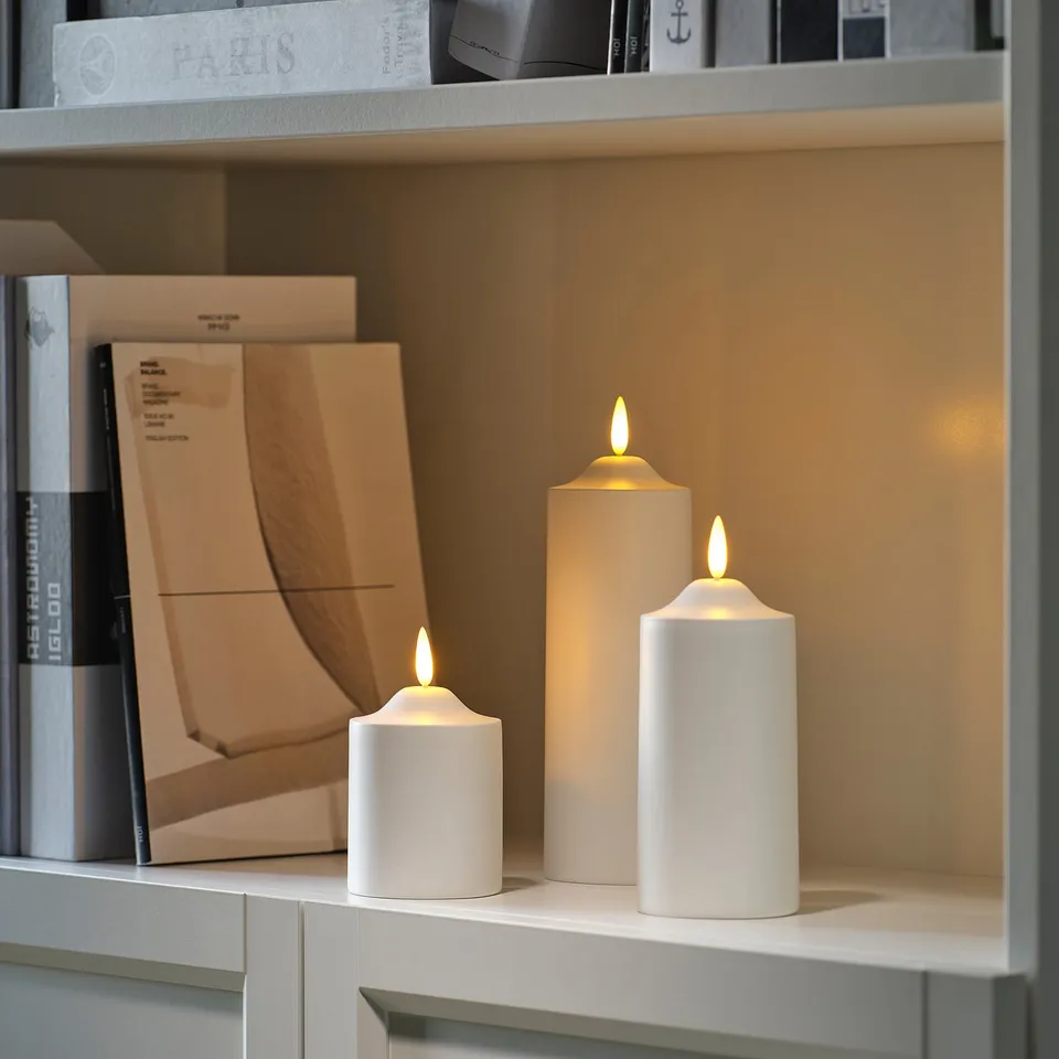 Three SMÅLOM white led candles of varying sizes on a shelf, surrounded by books and magazines.
