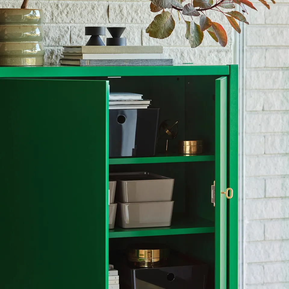 Green cabinet with brass lock, storing boxes and binders, against a white brick wall.