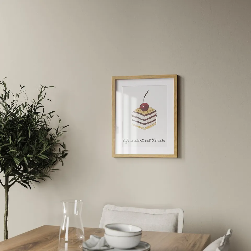 A cosy dining space with a beige framed print featuring a cake and cherry on the wall, beside a table with a glass carafe, cups, and a potted plant.