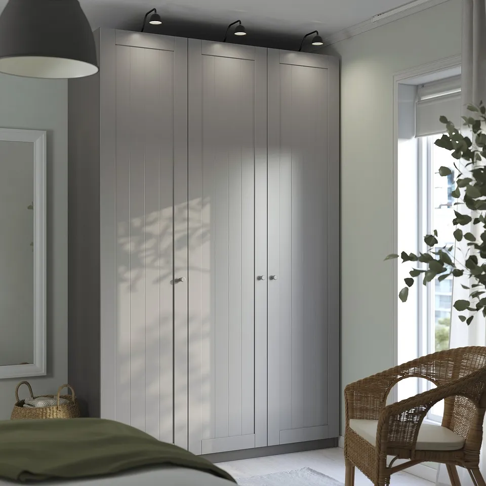 Modern bedroom with grey PAX wardrobe, lighting, and wicker chair by window.