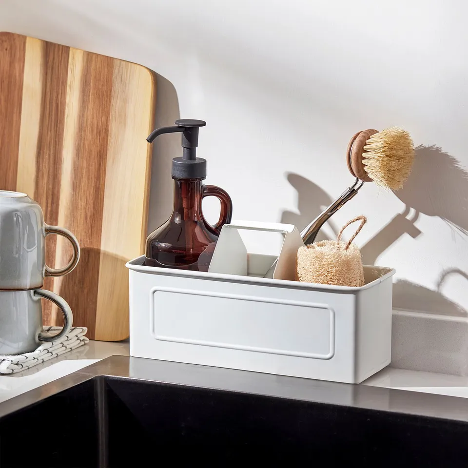 White kitchen caddy with handle, holding brown soap dispenser, brush, and sponge, organised near sink.
