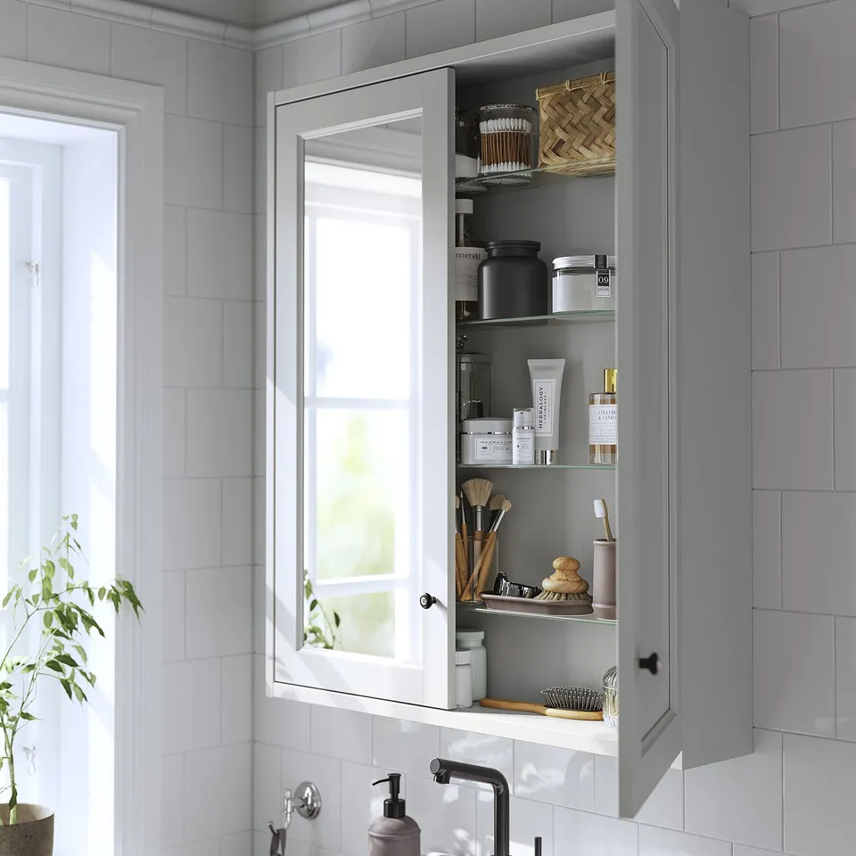 Open grey mirror cabinet displaying toiletries, mounted above white sink, set against tiled wall.