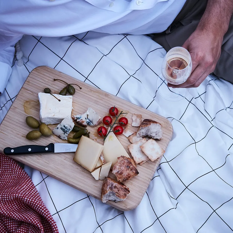A person in a white shirt partly visible holds a glass of wine. In front is a wooden cutting board with various cheeses, olives, and tomatoes.