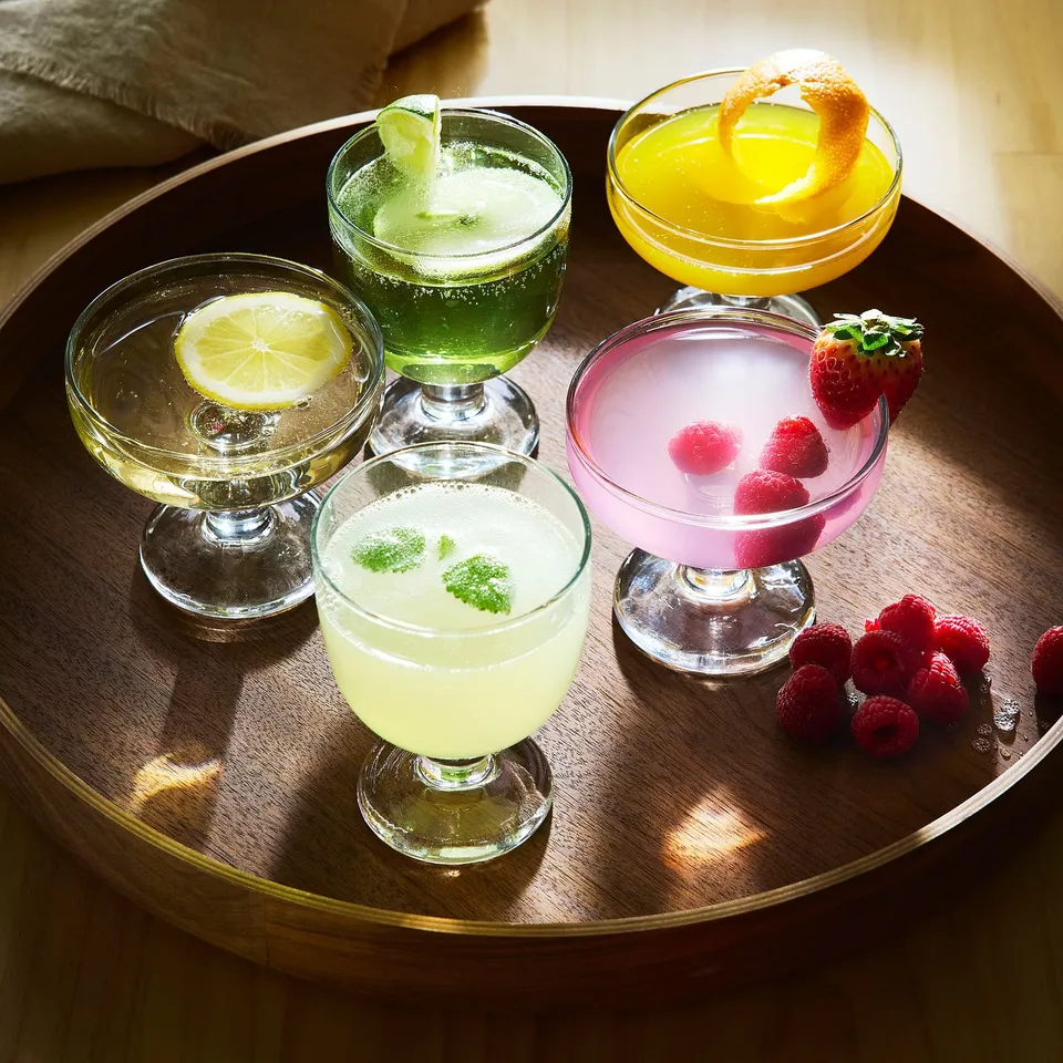Five colourful cocktails in goblet and coupe glasses, garnished with fruit slices, on a wooden tray, perfect for serving drinks.