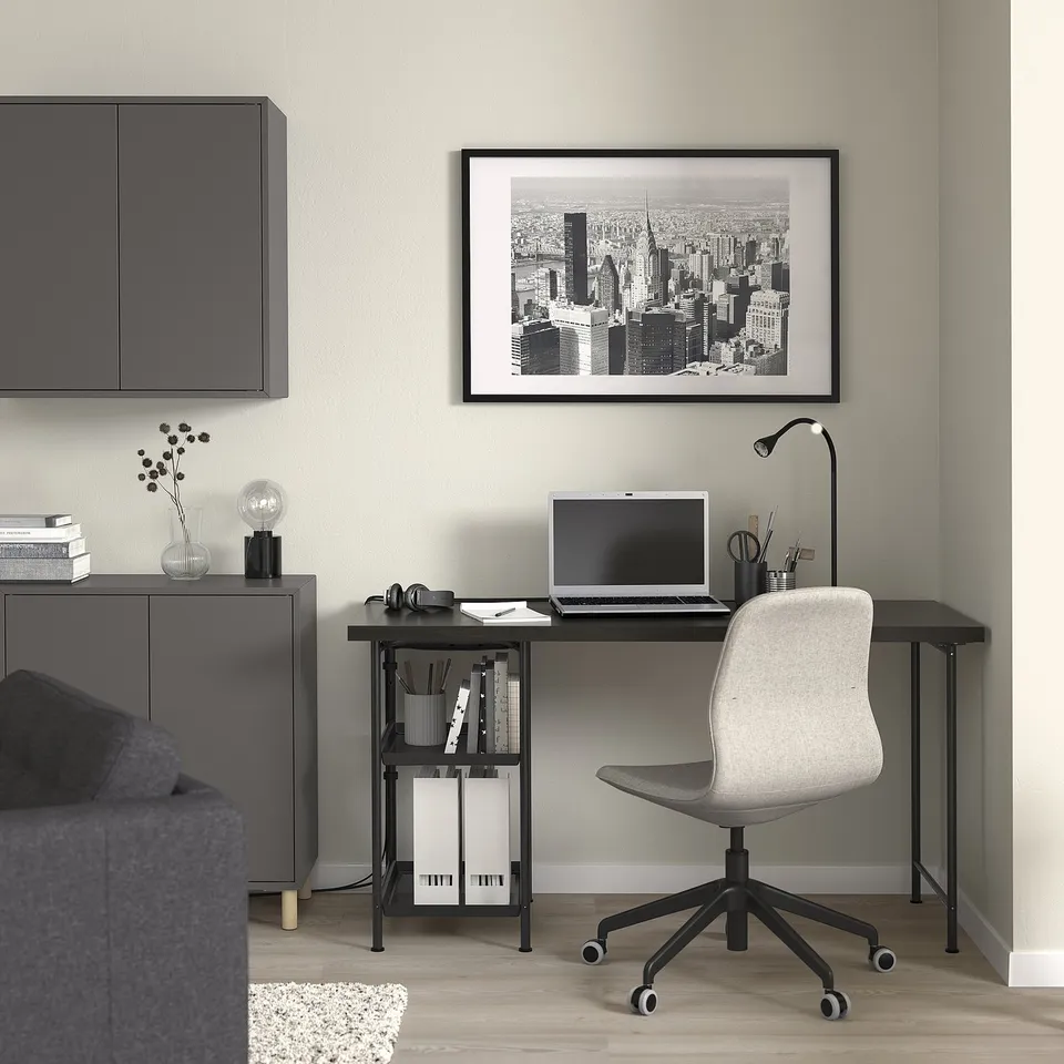Modern home office with SPÄND black metal desk, laptop, chair, and cityscape wall art. Desk has two shelves holding paper and books.