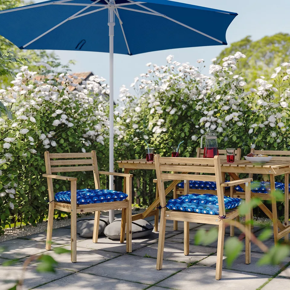 Outdoor patio set with blue sun umbrella, wooden table, and chairs with blue polka dot cushions, surrounded by white flowering bushes.