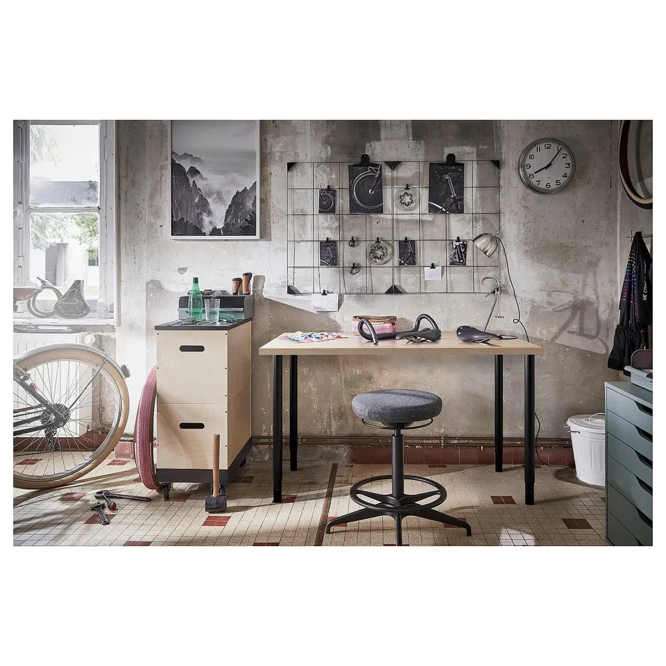 Industrial-style wooden desk with grey stool, wall clock, shelves, and bike.