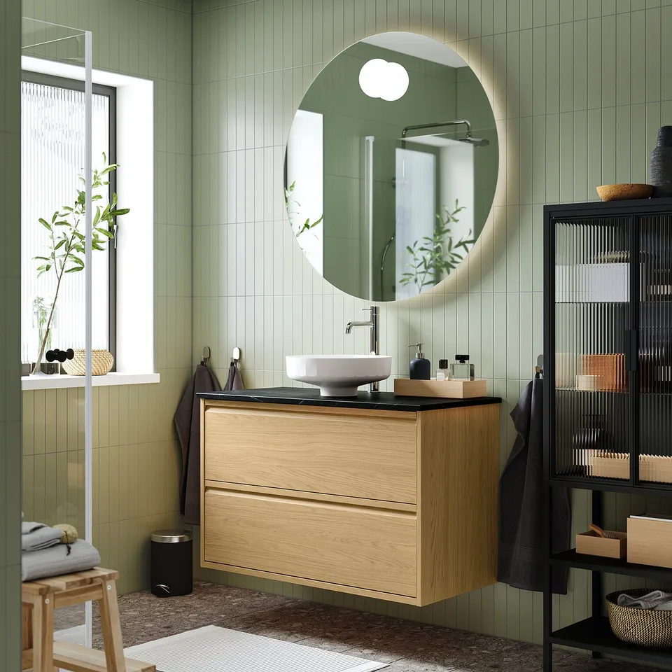 Modern bathroom with wood, green tiles, circular mirror, slim sink, decor on shelves.