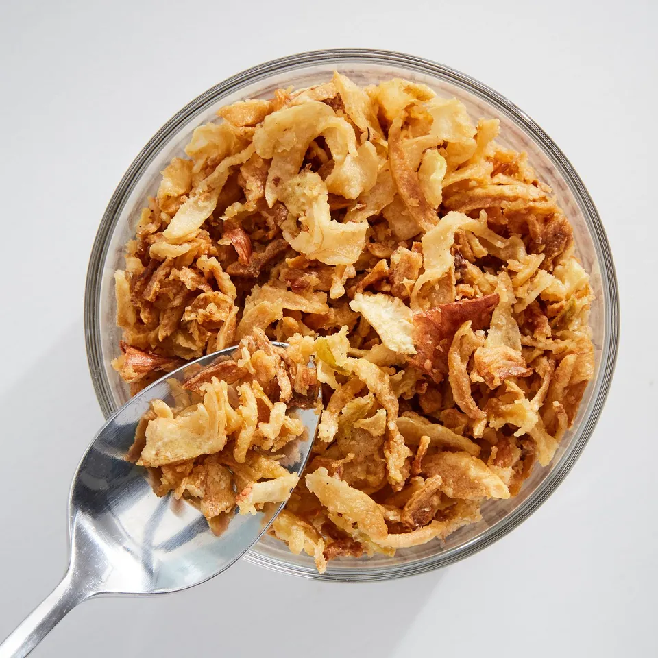 Glass bowl of crispy fried onions with spoon. Golden-brown, irregular shapes.