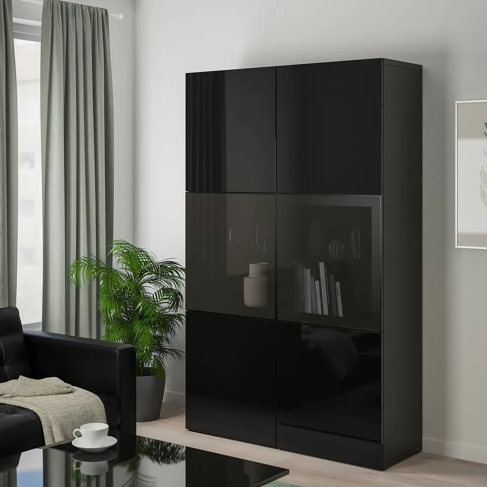 A black BESTÅ cabinet with glass doors, holding books and vases. It stands tall beside a window, near a dark chair and a plant.