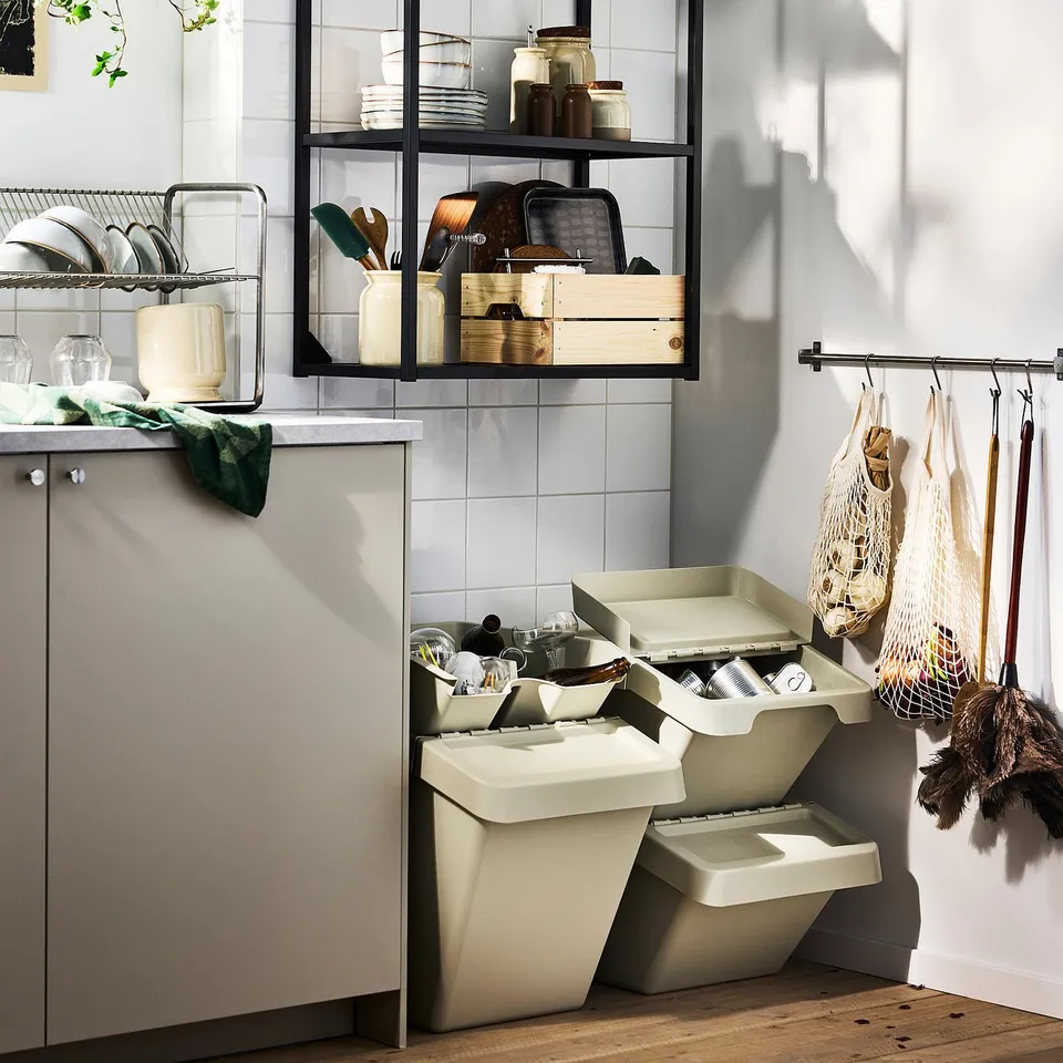 Modern kitchen with open shelves holding dishes, jars, and wooden boxes. Below, stacked beige recycling bins. Net bags hang with cleaning tools.