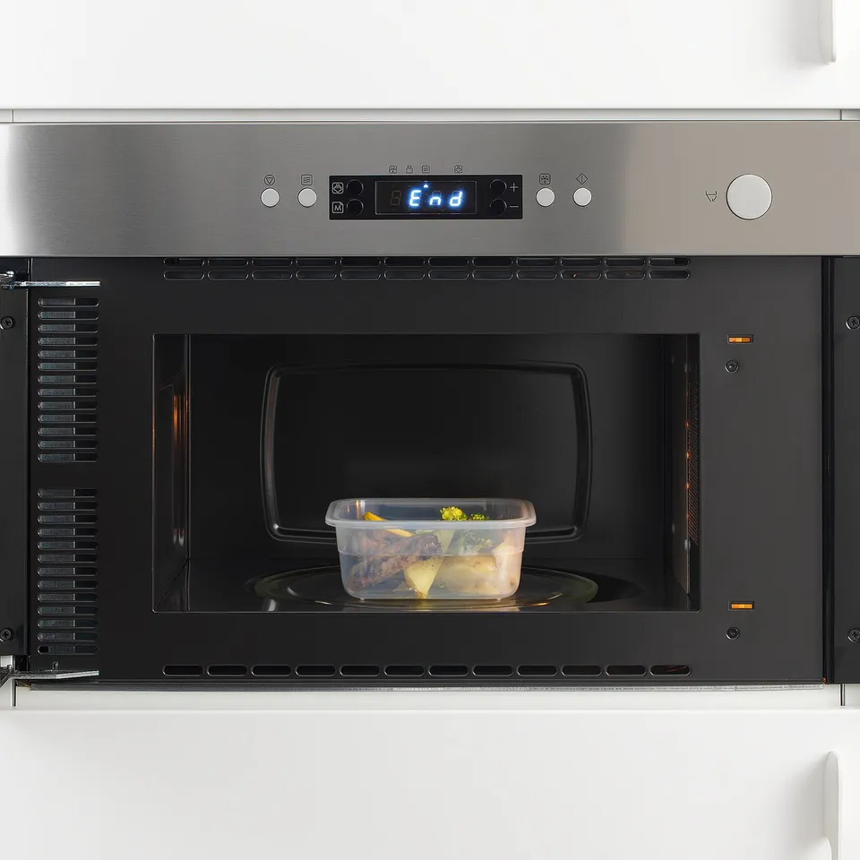 Stainless steel microwave with digital display and black interior, heating plastic container of food.