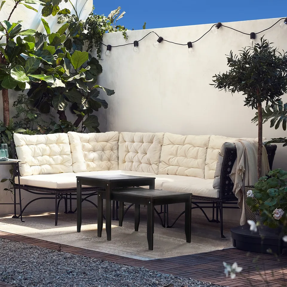 Modern patio with ÖRSKÄR wooden tables and white sectional sofa, surrounded by plants and string lights.