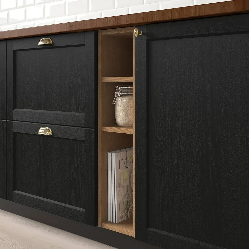 Black kitchen cabinet with open section displaying a jar and books. Golden handles, set against white brick wall.