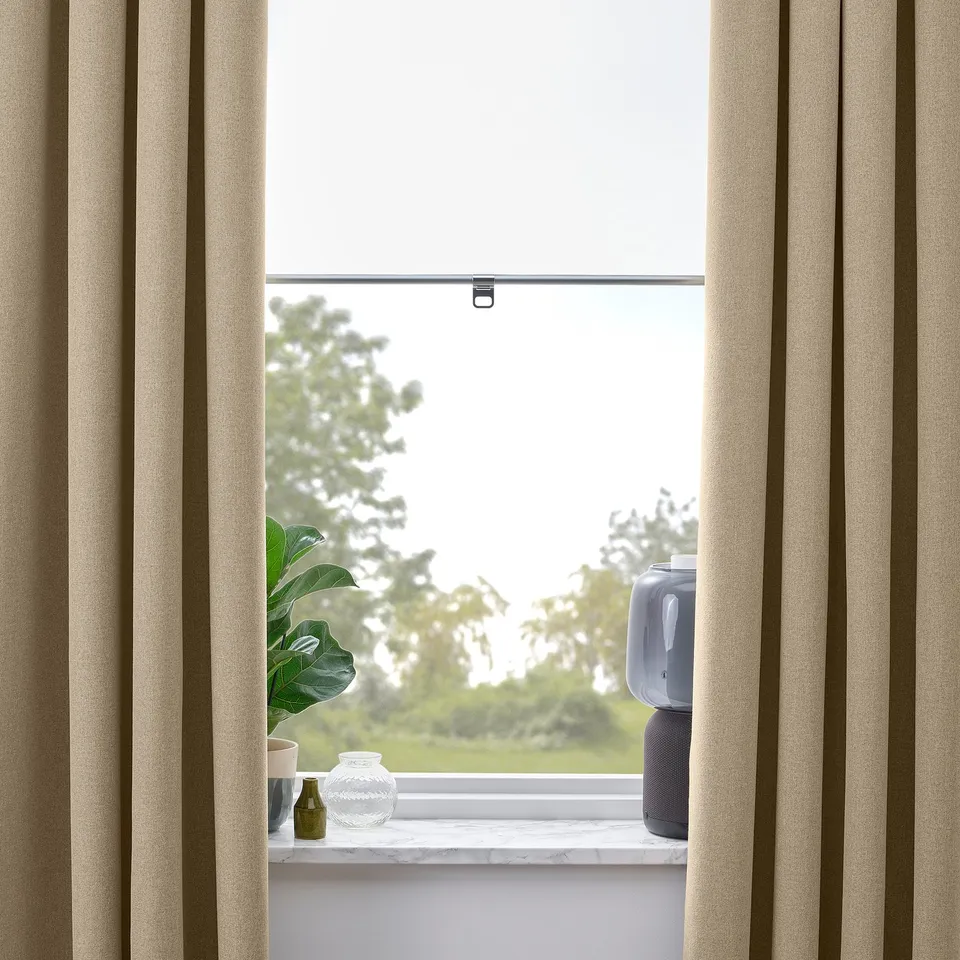 Window with yellow-beige room-darkening curtains partially open. Visible are a plant, vases, and an air purifier on the sill, framed by beige fabric.