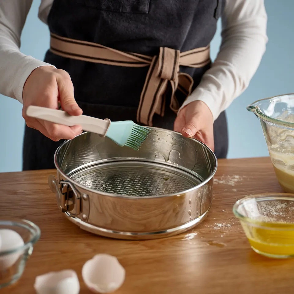 Person brushing oil on springform pan for baking. The pan is on a wooden table with ingredients nearby.