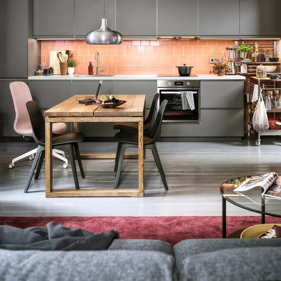 Modern kitchen with pink tiles, grey cabinets, wooden table, and hanging pendant lights.