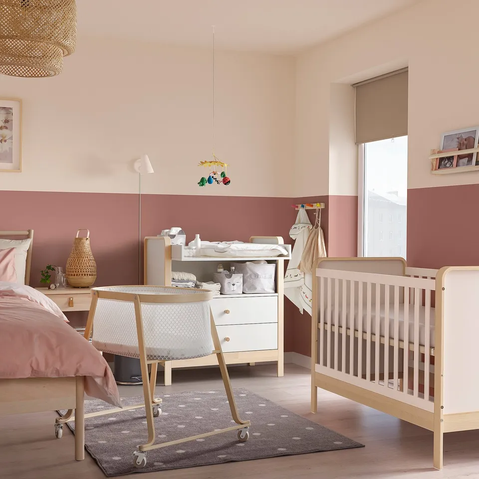 Scandinavian baby room with bassinet, changing table, crib, and pink-beige walls.