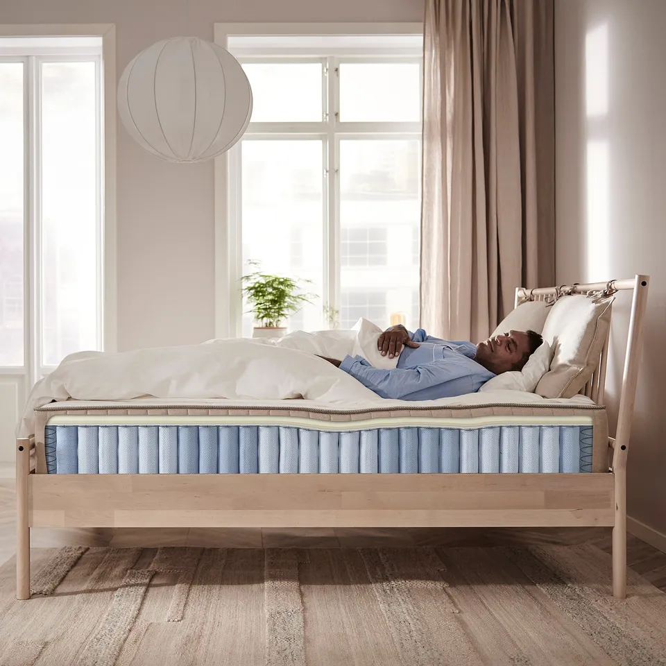Person resting on firm blue mattress in modern bedroom with large windows and light wood bed frame.