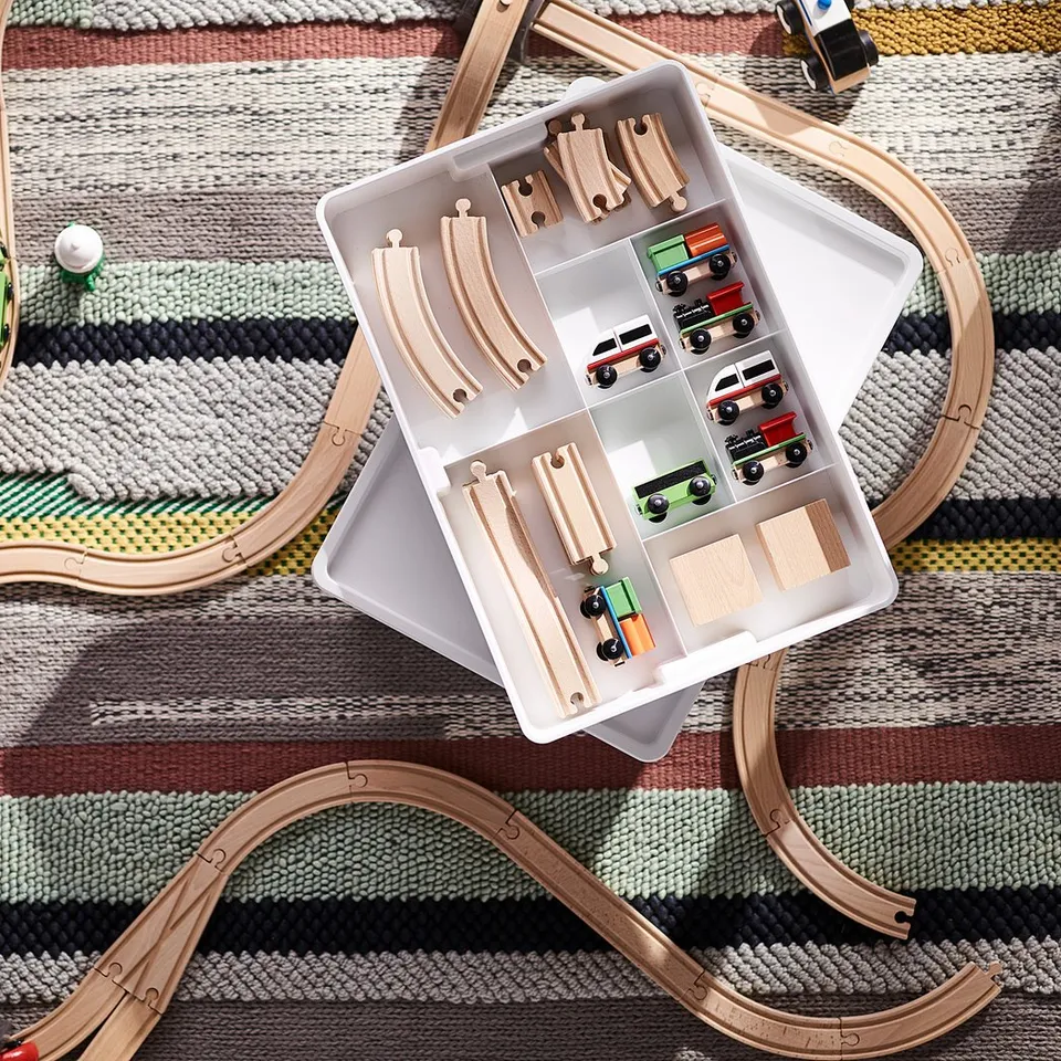 Wooden toy train set with tracks, cars, and storage box on a patterned rug.